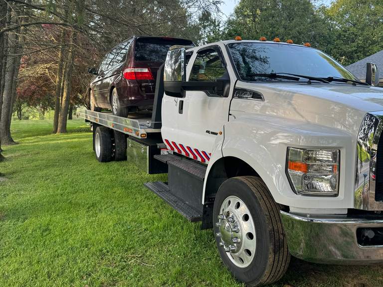 White flatbed tow truck carrying a maroon Honda Odyssey minivan, parked on a grassy lawn