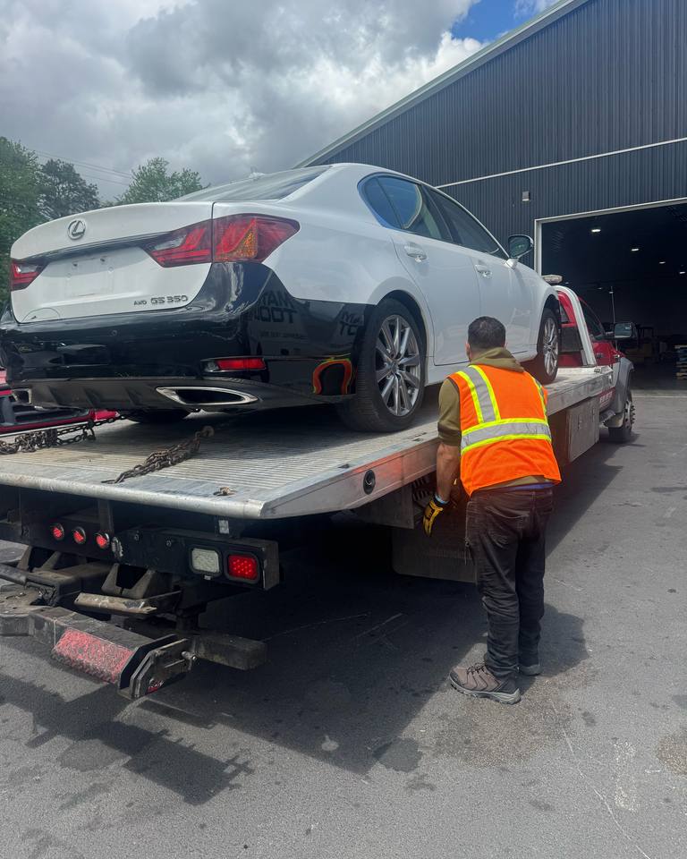 Worker in a safety vest securing a white Lexus GS 350 on a flatbed outside a warehouse
