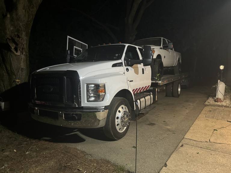 White Ford F-650 flatbed carrying a white pickup truck on a residential driveway at night